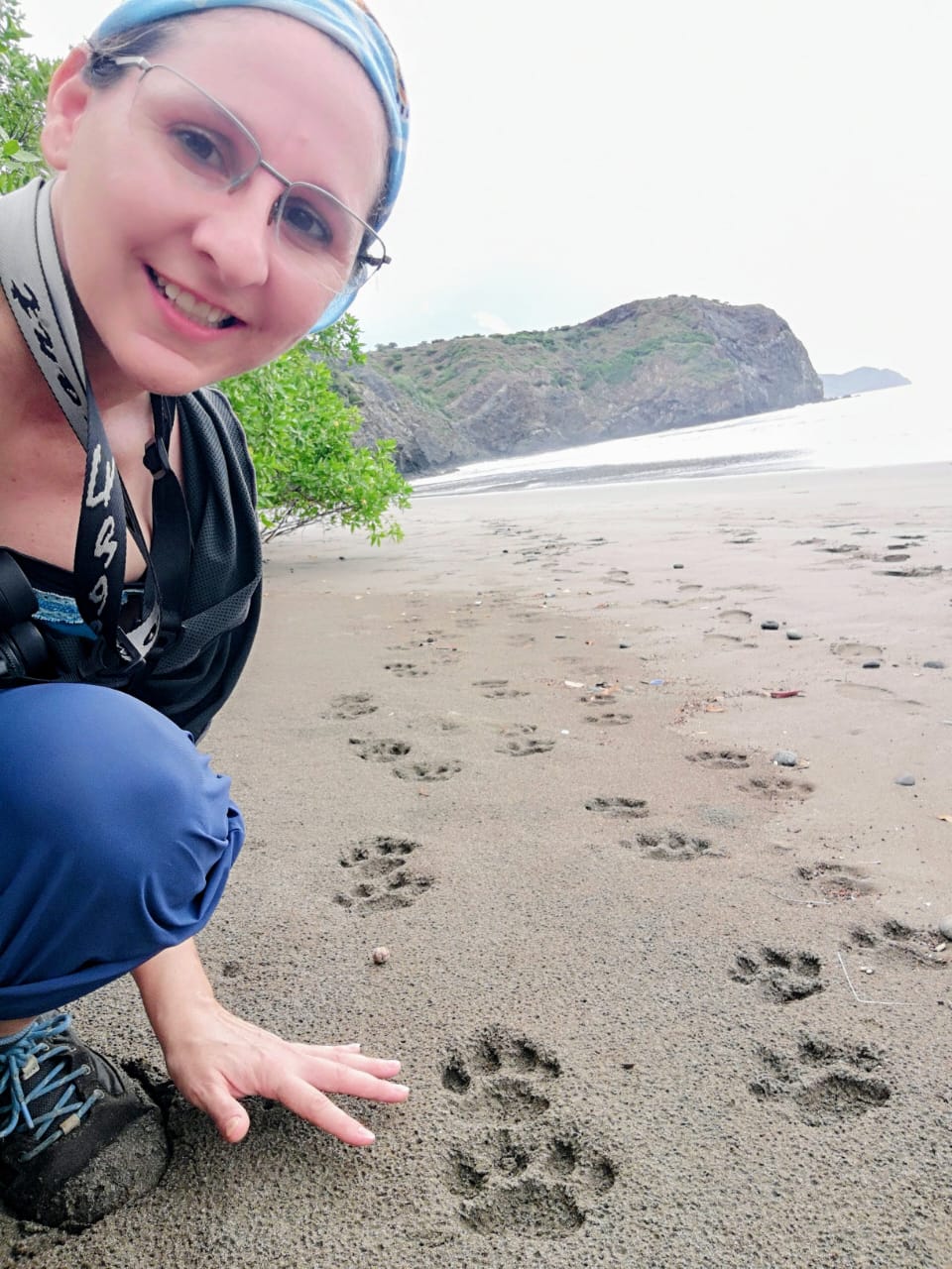 Ronit Amit headshot, crouching down next to some big paw prints on a beach